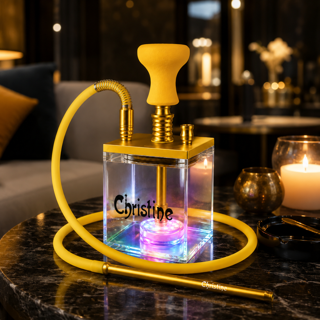 A transparent acrylic hookah-style water pipe with a yellow mouthpiece, yellow hose, and gold-tone metal components sits on a dark marble table in a dimly lit lounge setting. The front of the clear box is printed with the name &