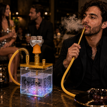 A man in a black suit smokes a hookah with a yellow hose and gold metal components. The hookah has a clear square base filled with glowing blue and purple LED lights, with the name &