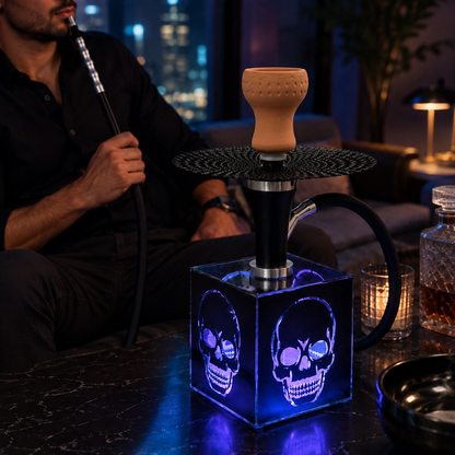 A skull-themed hookah set on a dark marble table in a dimly lit lounge. The hookah has a glowing blue LED cube base with skull designs, a black metal stem, and a beige ceramic bowl.