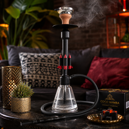A black hookah set with a clear glass base, black hose, and metallic bowl, displayed on a dark table in a cozy lounge setting with a visible COCO HAMRA box and lit shisha joints.