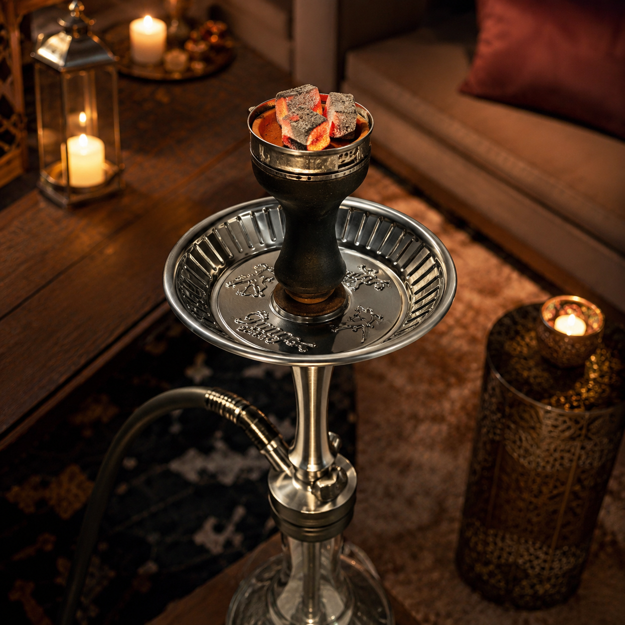 A polished silver and black hookah set with glowing coals in the bowl, placed on a wooden table in a warmly lit lounge setting with candles and cushions.