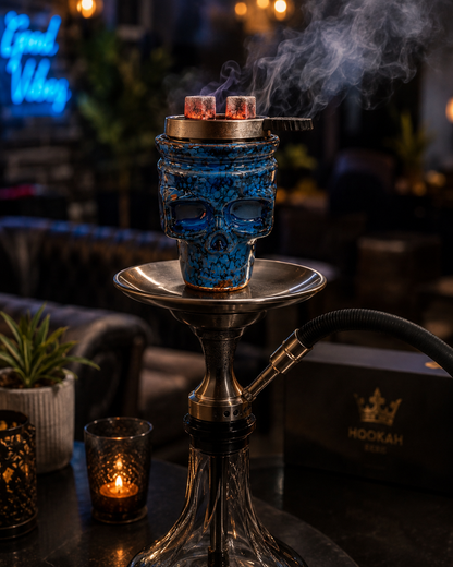 A skull-shaped hookah with a textured blue and black pattern sits on a silver metal stand on a dark table, with smoke rising from the burning fuel at the top. A lit candle glows nearby, and a black box with a gold crown logo and the word &